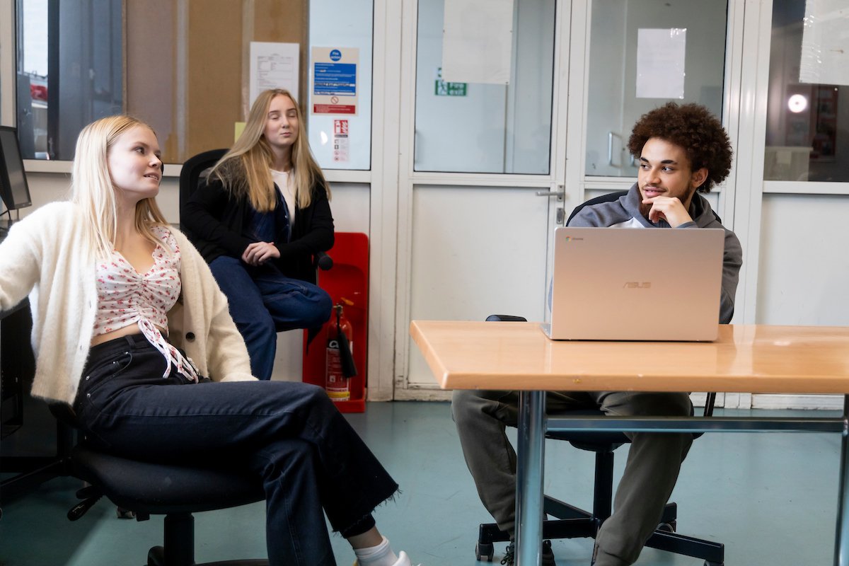 2 young women and 1 young man are sitting around a desk. The young man has his laptop open. They are researching 2CB and 2C-i drugs. This is a full-body image.