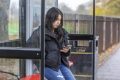 A young person is sitting at a bus stop wearing a black jacket. She is on her phone looking up how to put a tampon in correctly