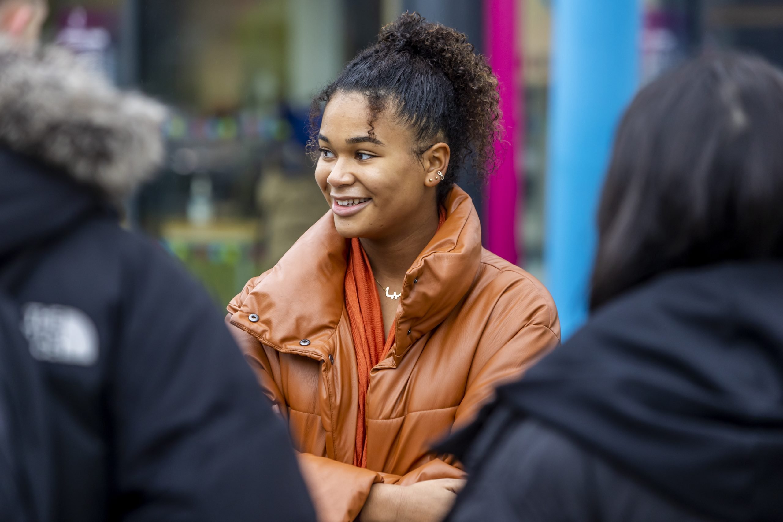 A young person is standing outside wearing an orange coat and smiling. They are thinking about when to come off antidepressants