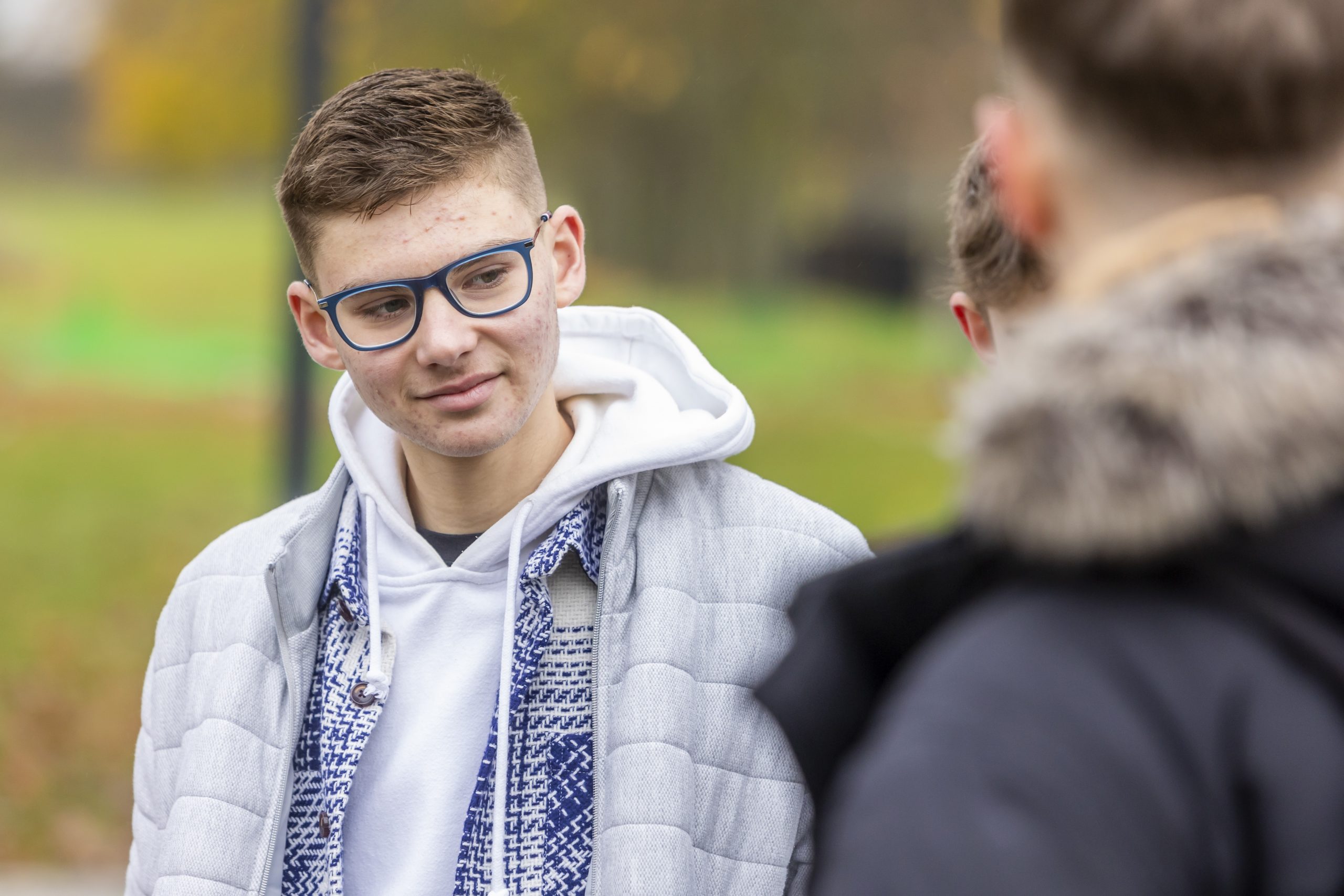 Two young people are standing outside talking about Gonorrhoea (aka, the clap) STI. One is wearing a white jacket and glasses and the other is facing away wearing a black coat.