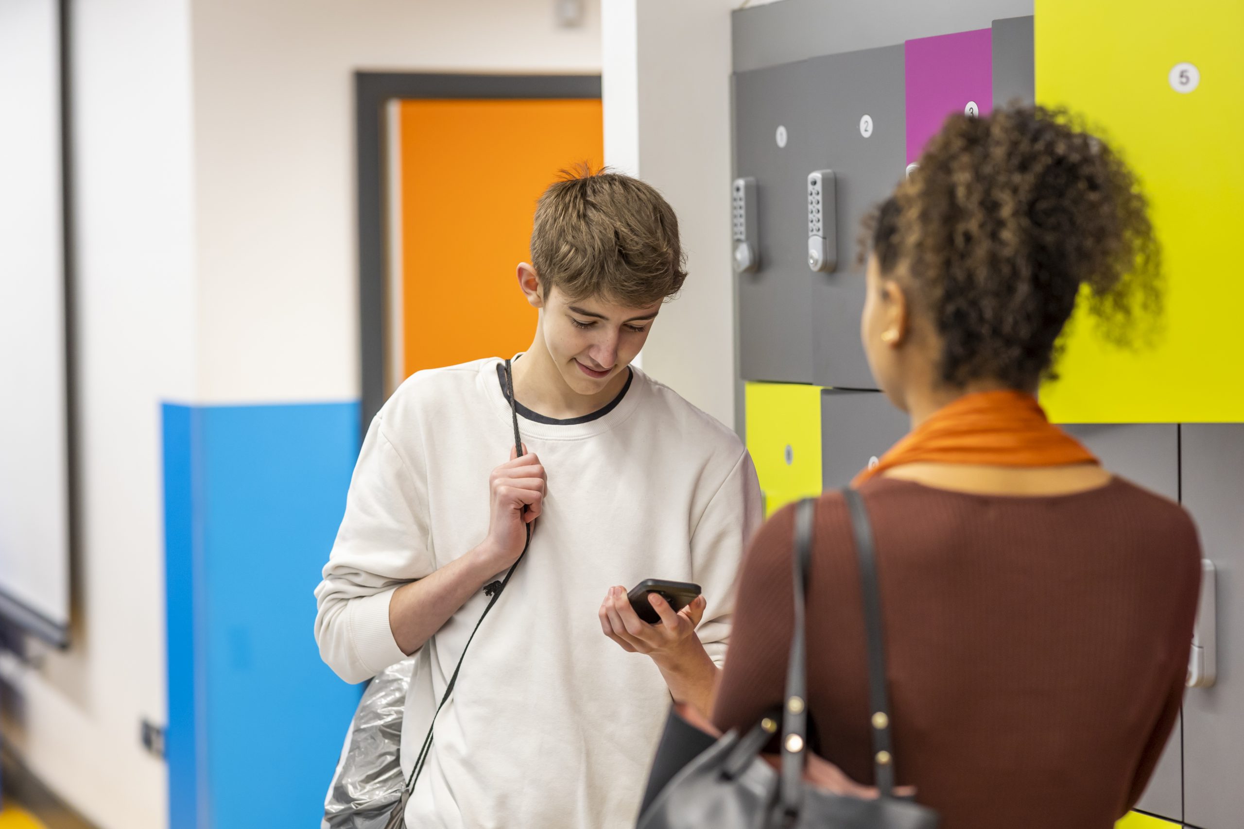 Two young people are talking in a school corridor, one is looking at their phone googling, "what is hepatitis c?"