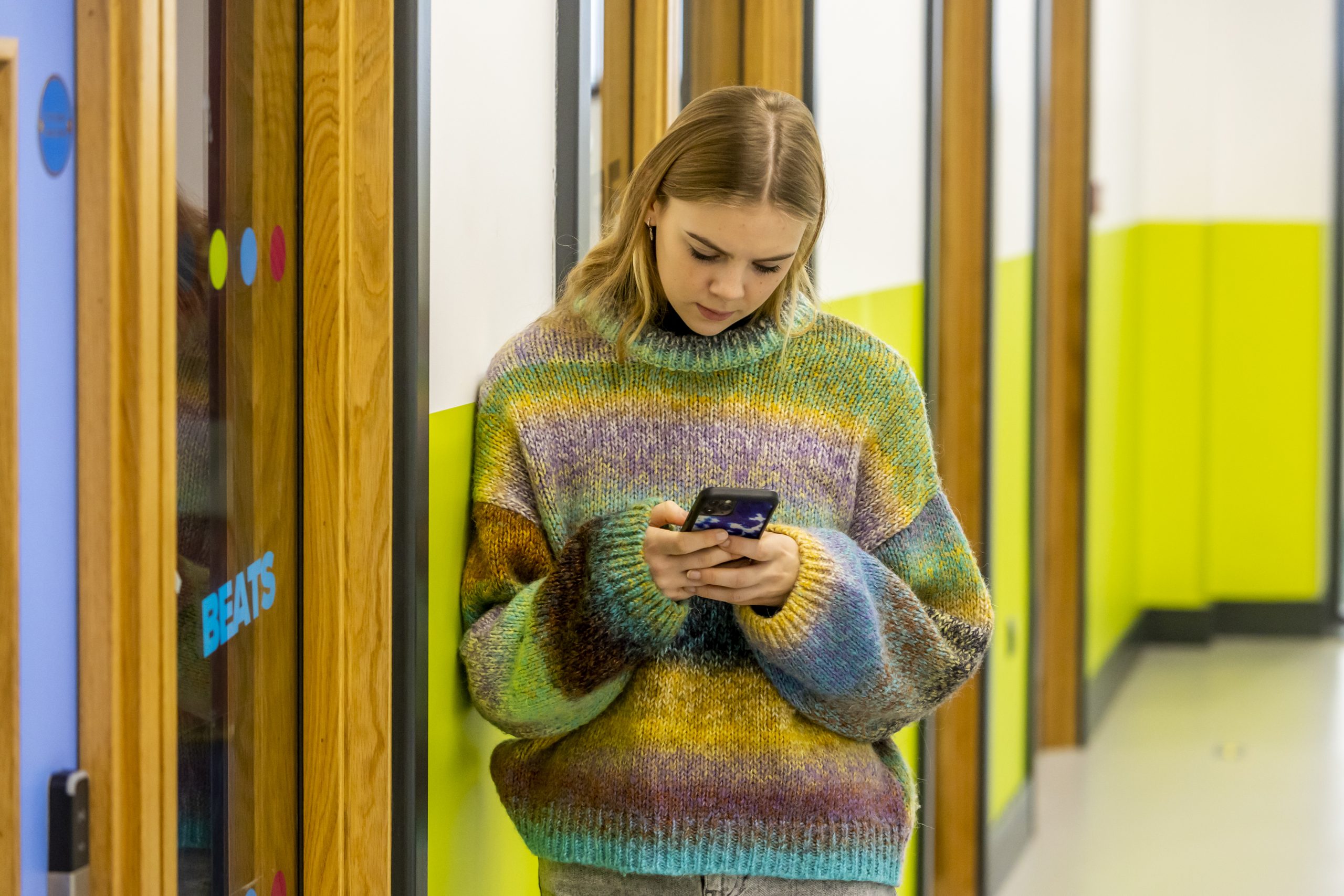 A young person is standing in a corridor wearing a rainbow striped jumper. They are looking at their phone to find out how to know if you've got hepatitis B