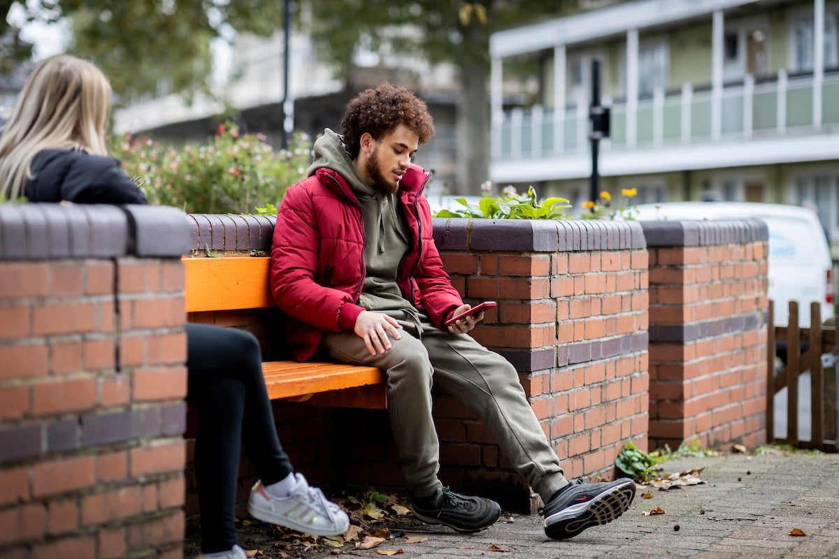 A young man is sitting on a bench. He is looking up Rohypnol. This is a full-body image.