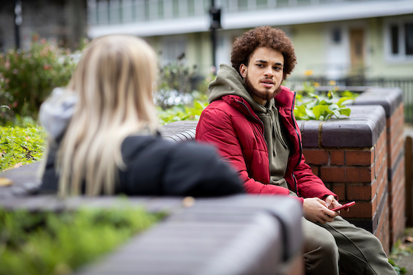 A young person is sitting on a bench wearing a red coat, talking to another young person about men's domestic violence