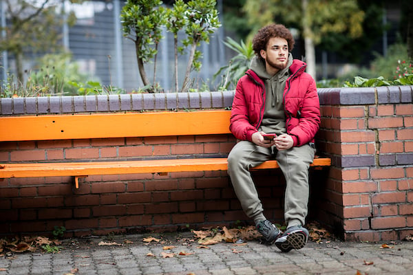 A young person is sitting outside on a bench wearing a red jacket. They are thinking about why they have aching testicles