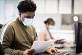 A young man is sitting on a desk. He is wearing a mask. He is looking at a paper on DMT. This is a wide-angle image.