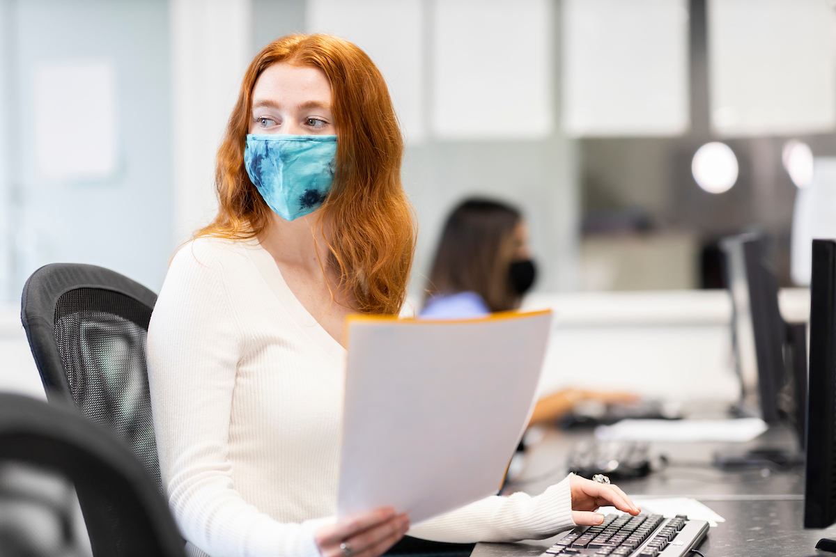 A young woman is sitting at a desk. She is wearing a mask. She is reading about mephedrone. This is a wide-angle image.