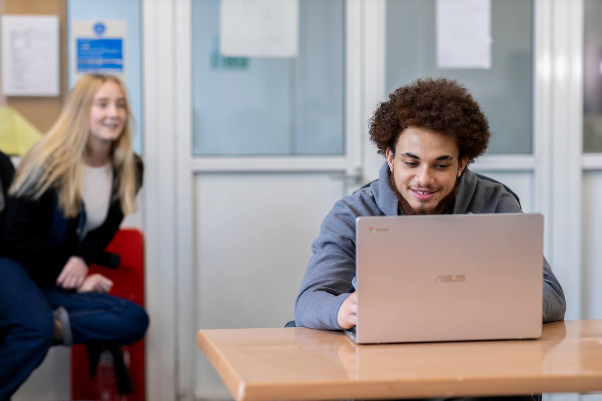 A young person is on their laptop laughing with their friend, who sits behind them