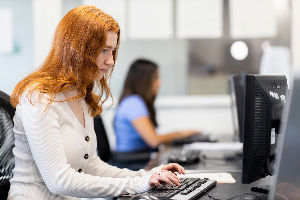 A young, red headed woman is sitting at a desktop. She is researching acid (LSD). This is a wide-angle image.