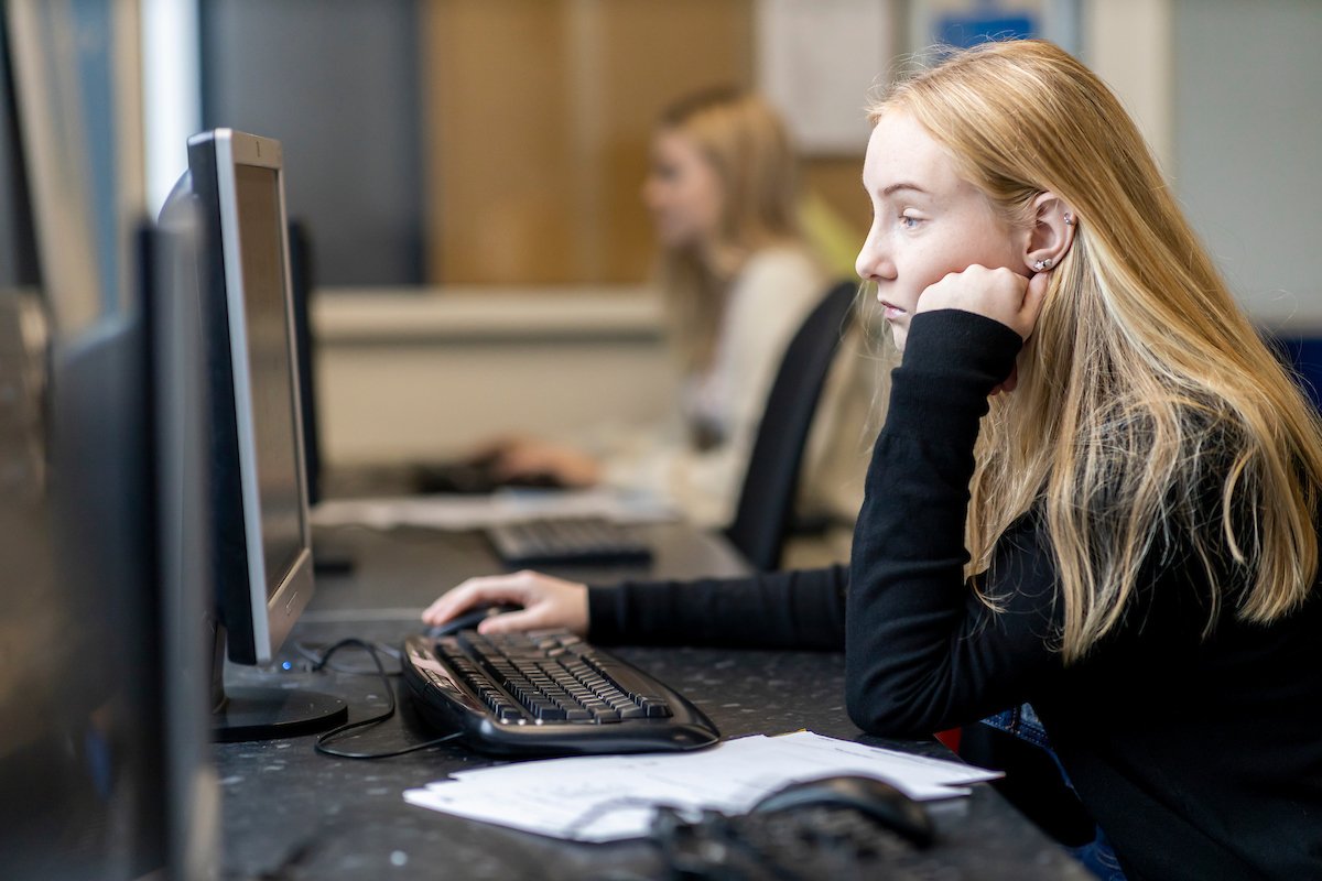 A young, blonde woman is sitting at a desktop. She has her hand on her fact. She is looking up Morning Glory. This is a wide-angle image.