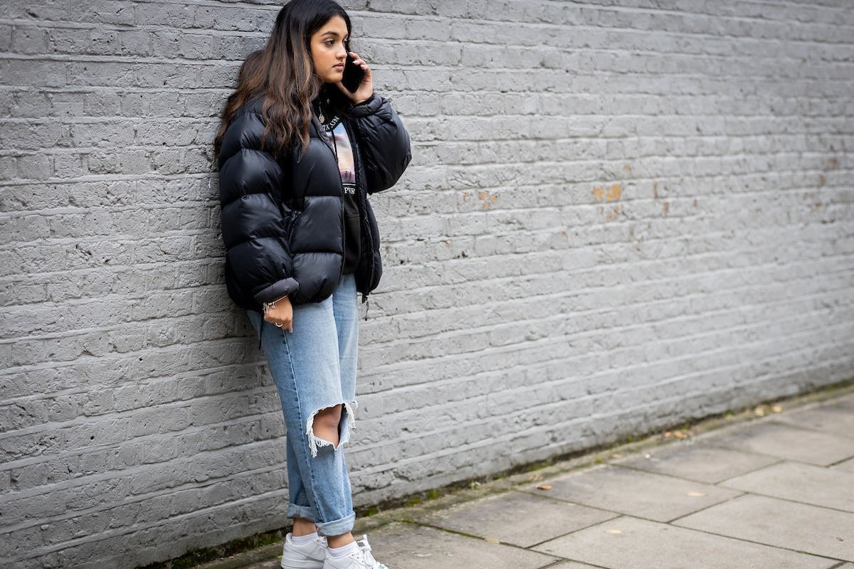 A young woman is standing in an alley calling an addiction helpline. She is wearing a black puffer jackt, black shirt and ripped jeans. It is a full-body image.
