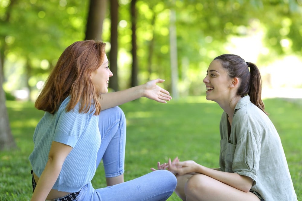 Two young women are in a park. They are discussing body odour. This is a wide-angle image.