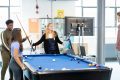 A group of young people are standing around a pool table. They are planning a cocktail party. They look happy. This is a full-body image.