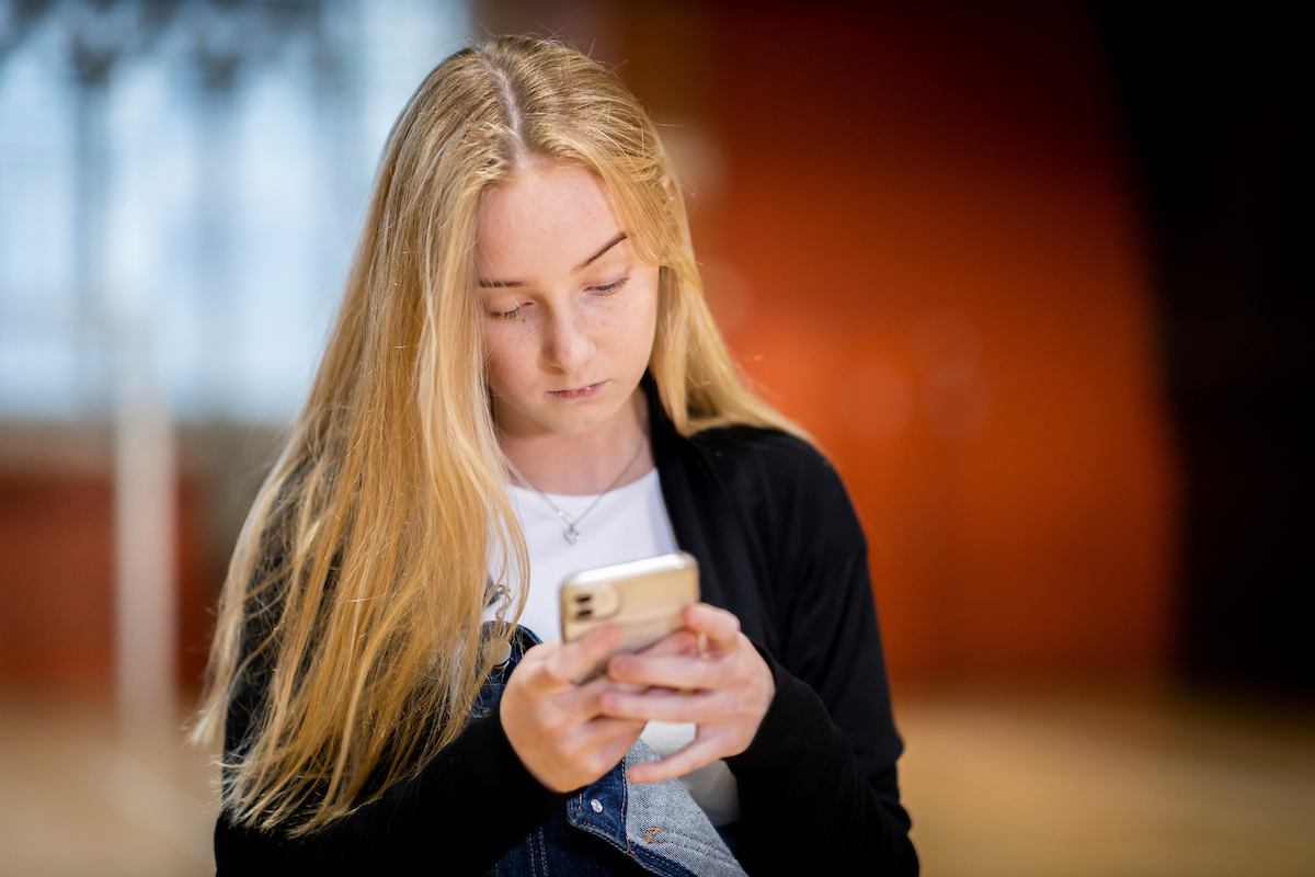 A young, blonde woman is staring at her phone. She is wearing a black cardigan and white top. She is looking up cocaine. This is a close-up image