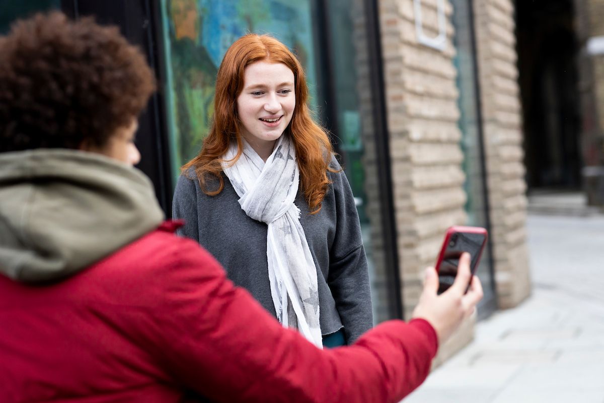 A young red-headed woman is looking at a picture on her friend's phone. It explains the rules to classic drinking games. The young man has his back facing the camera. This is a wide-angle image.
