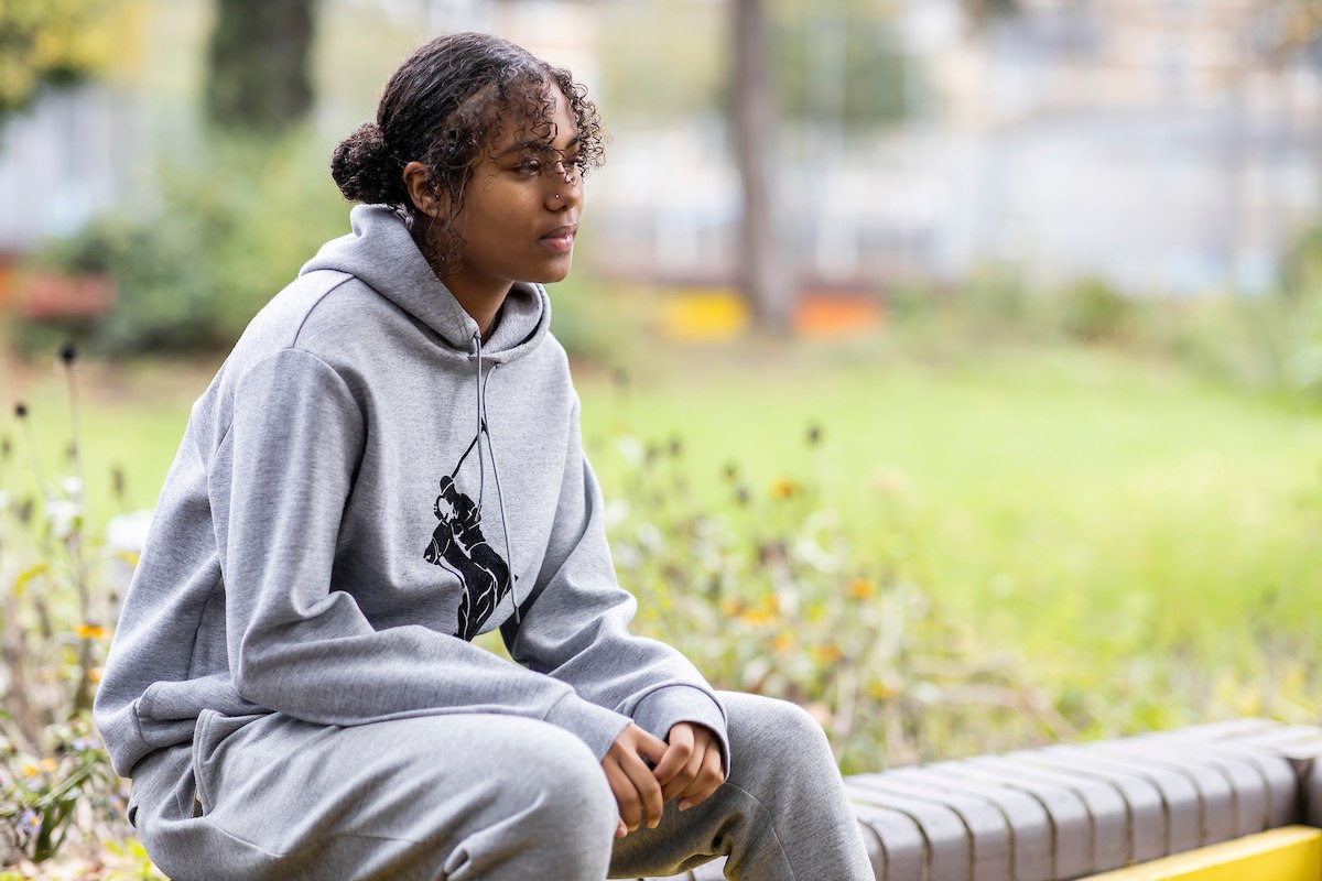 A young woman is sitting on a bench alone. She is wearing a grey sweatset. She is thinking about drugs rape. This is a wide-angle image.