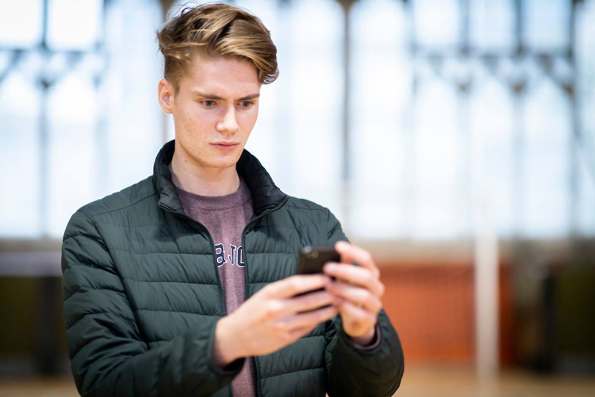 A young man is staring at his phone worriedly. He has seen a picture of one of his embarrassing drunk moments. He is wearing a puffer jacket and a purple t-shirt. This is a close-up image.