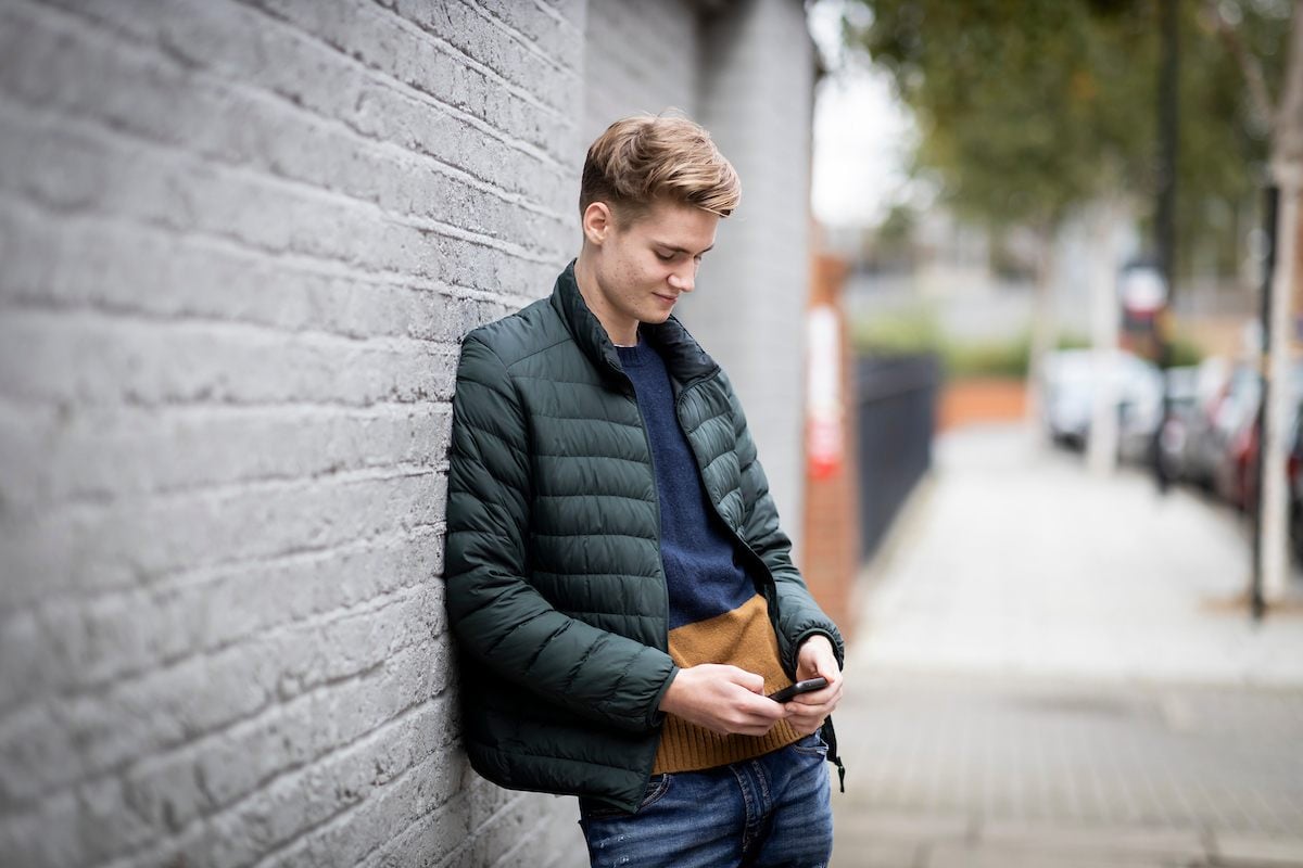A young man is leaning on a wall, staring at his phone. He is looking up ephedrine. He is wearing a puffer jacket. This is a wide-angle image.