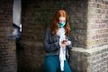 A red-headed young woman is leaning on a brick wall. She is looking up how to get help for addiction. She is wearing a white scarf, a grey zip-up and a blue mask. The photo is cut-off at her waist.