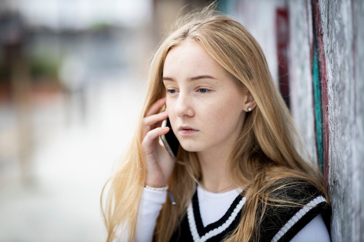 A young, blonde woman is on the phone. She is wearing a long-sleeved white shirt with a black vest on top. She is finding out how to help an addict. She looks upset. This is a close-up image.