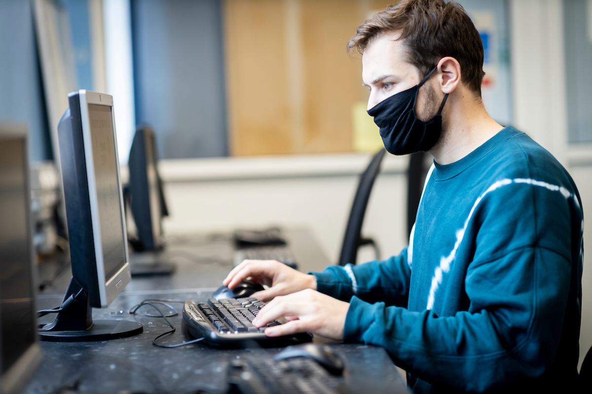 A young man is sitting at a desktop. He is looking up how drugs get into the UK. He is concentrating on the research. This is a wide-angle image.