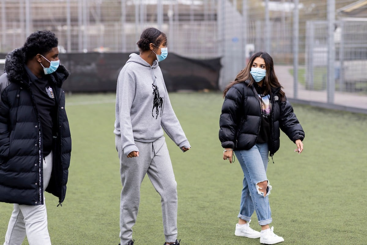 Three young people are walking on a field. They are wearing masks. They are discussing their drunk friend from last night. This is a wide-angle image.