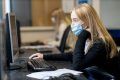 A young, blonde woman is sitting at a desktop. She is wearing a mask. She is researching Crystal Meth. This is a wide-angle image.