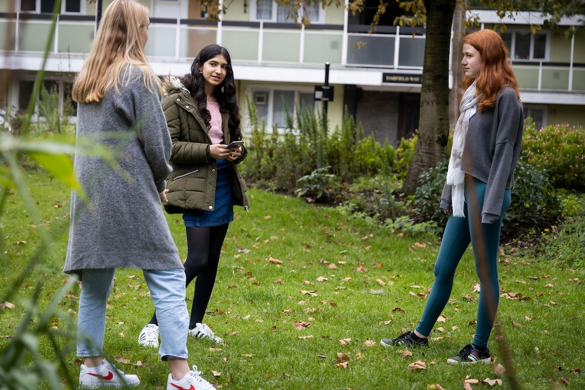 Three young people are standing on some grass. They are in a semi-circle. One of the young women looks worried because she can't afford to go out. This is a full-body image.
