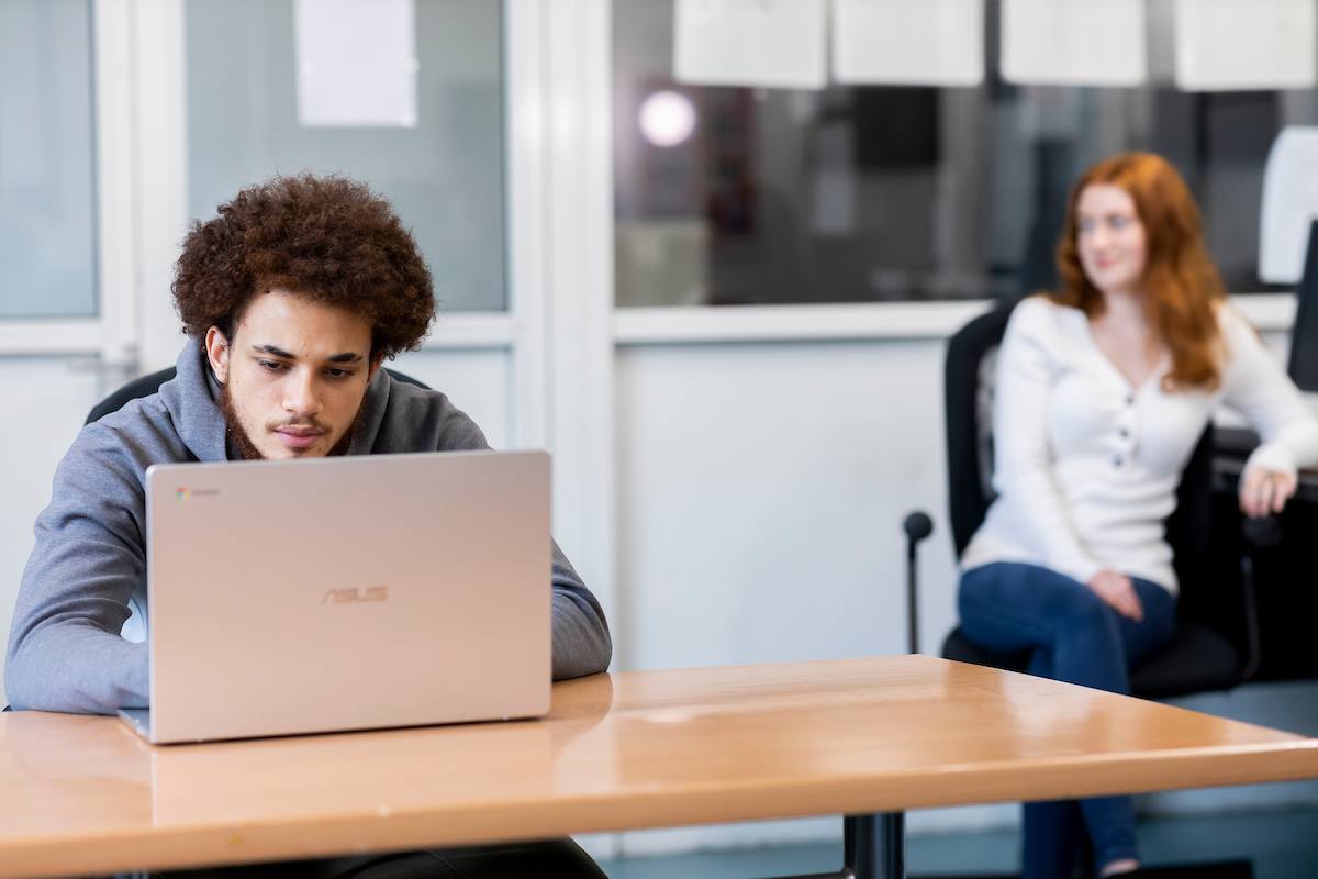 A young man is sitting at his laptop. He wants to buy prescription drugs online. A young red-headed woman is sitting behind him. This is a wide-angle image.