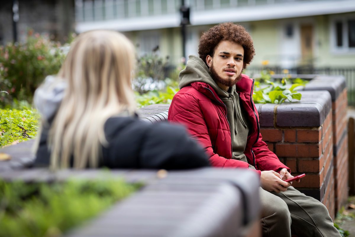 A young man is sitting on a bench. He is talking to his friend about his overdose. She is a young, blonde woman. Her back is facing the camera. This is a wide-angle image.