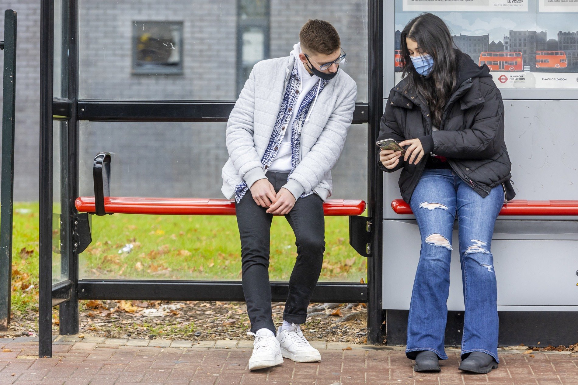 Two young people are at a bus stop. They are looking up PAYE tax. This is a full-body image.