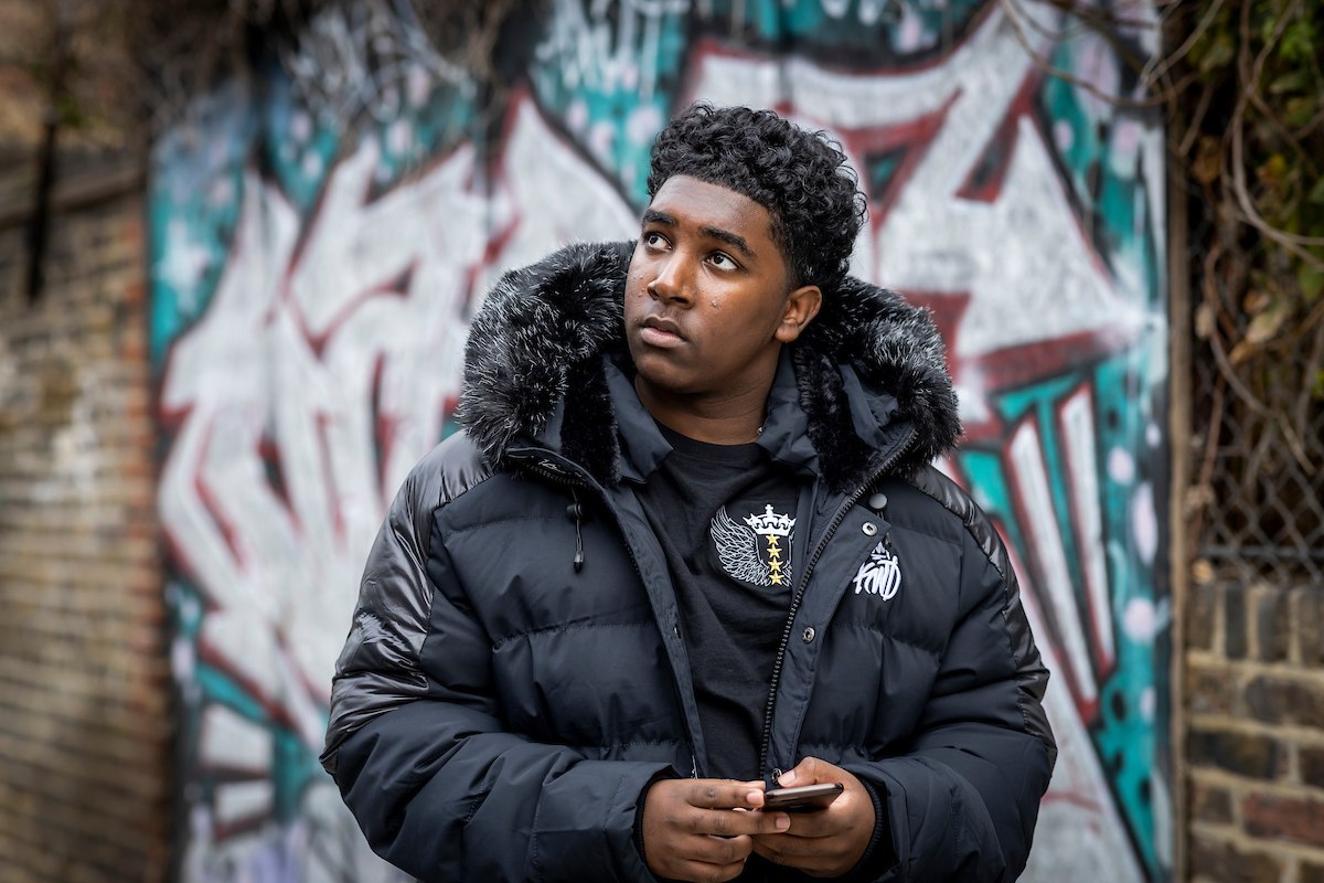 A young man is standing in front of graffiti. He is wearing a black puffer jacket and black shirt. He has just had a drugs relapse. He is looking off into the distance. This is a close-up image