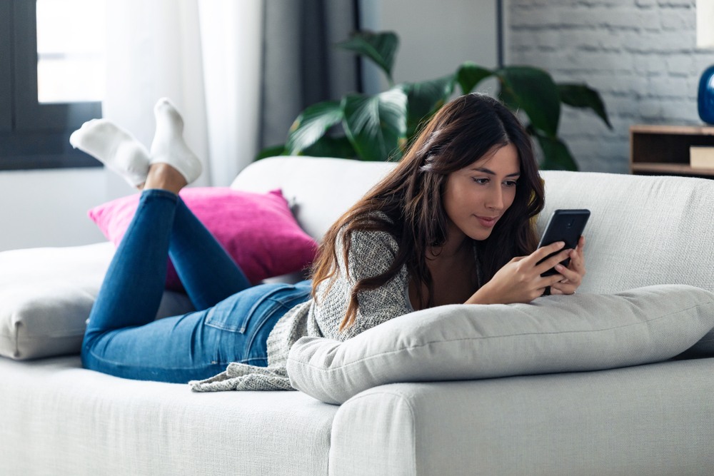 A young woman is sitting on the sofa with her phone. She is thinking about sex and self-esteem. This is a wide-angle image.