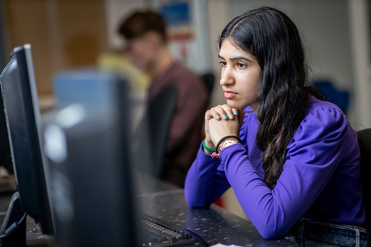 A young woman is sitting at a desktop. She is holding her fists together, in thought. She is researching solvent abuse. This is a wide-angle image.