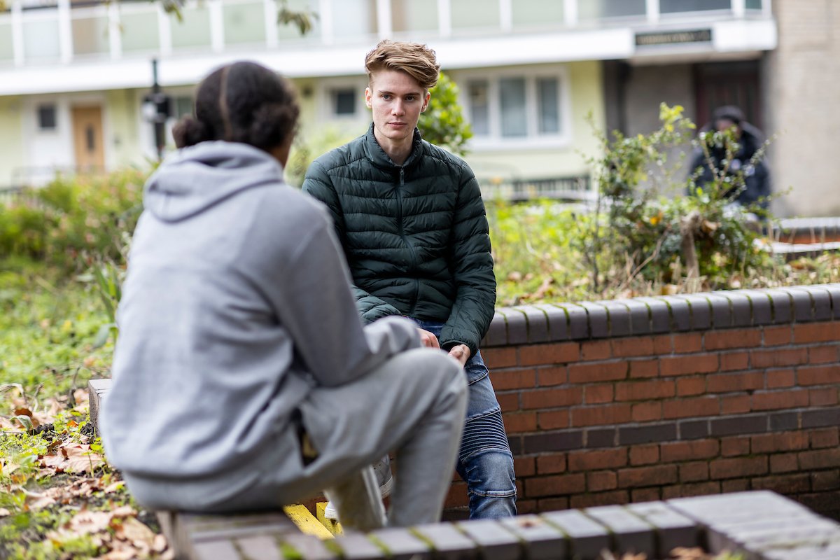 A young man is talking to a young woman about staying off drugs. They are sitting on a brick wall. He is wearing a zipup and looks upset. She is facing him, wearing a grey hoodie. This is a wide-angle image.