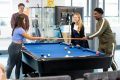 A group of young people are standing around a pool table. A young man and woman are hitting their pool sticks together. They are enjoying life being teetotal. This is a wide-angle image.