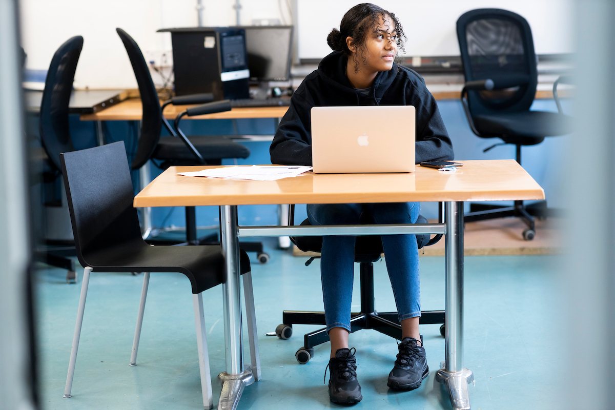 A young woman is sitting at a desk on her laptop. She is looking up drug addiction treatments. She is wearing a black hoodie and blue jeans. She is looking sideways nervously. This is a full-body image.