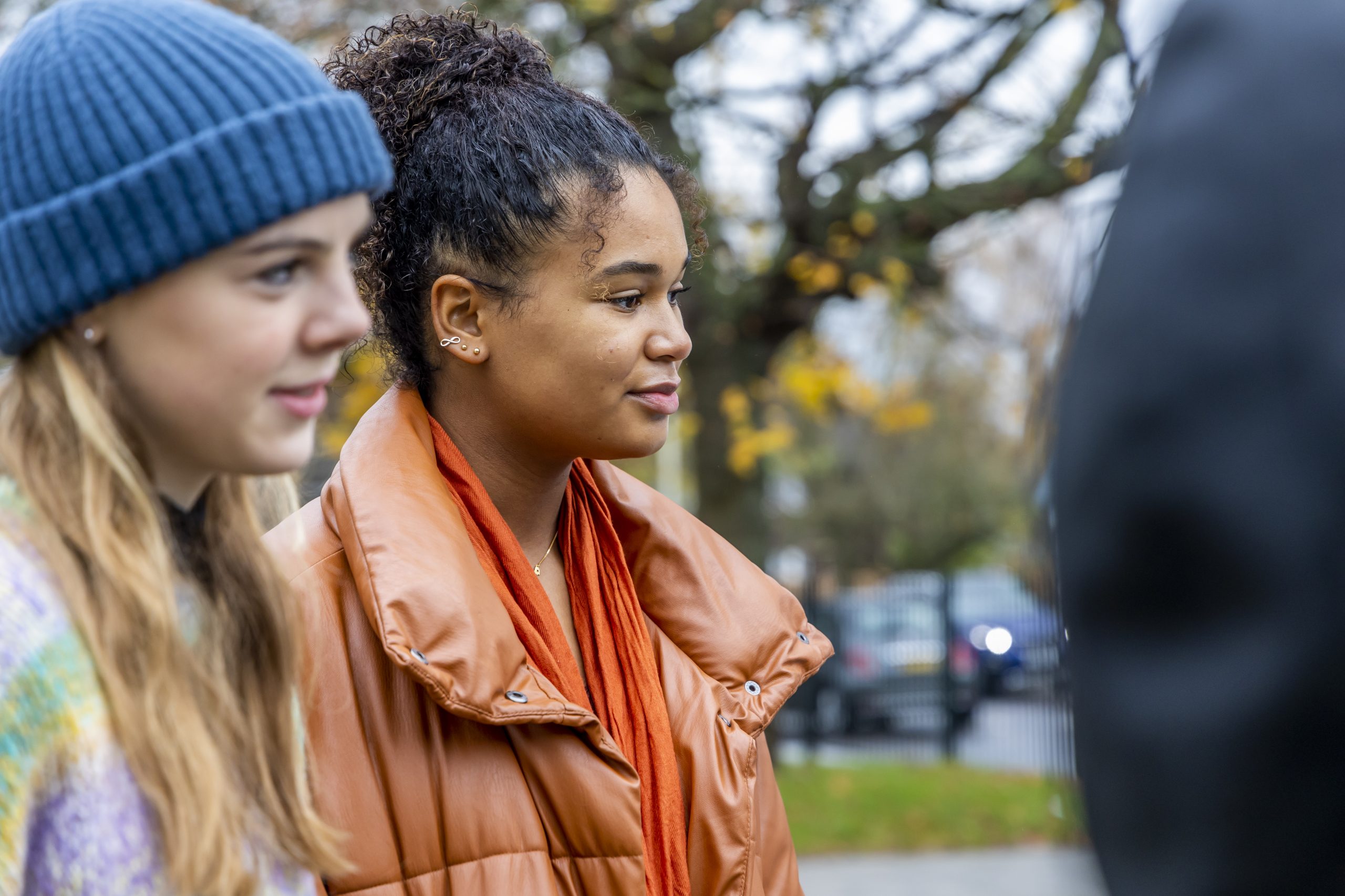 Two young people are standing outside talking about how to help a partner with anxiety. One is wearing an orange jacket and the other a rainbow jumper and a blue top