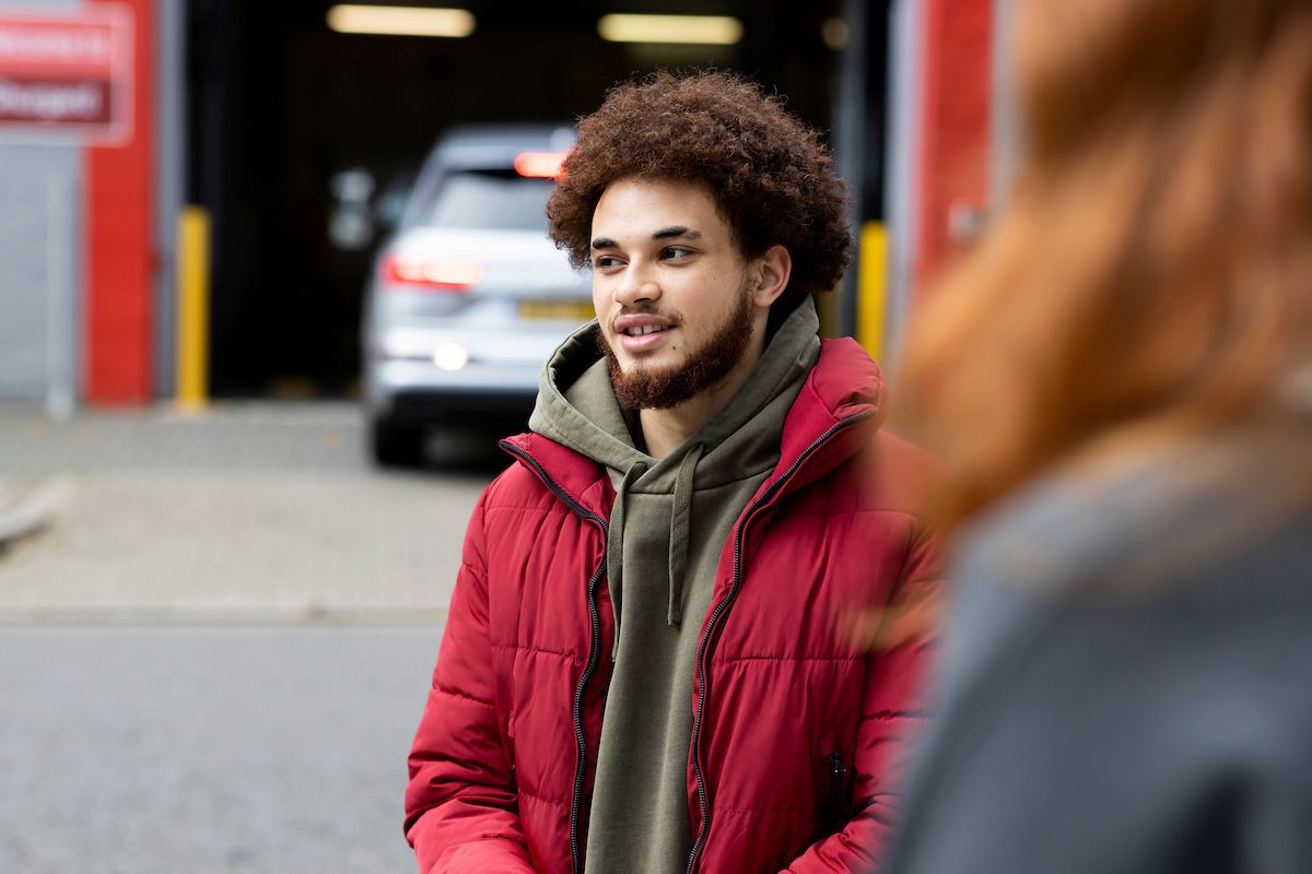 A young man in a red jacket is standing in front of a parked car talking to a friend. He is saying, "I hate my life" and asking for advice.