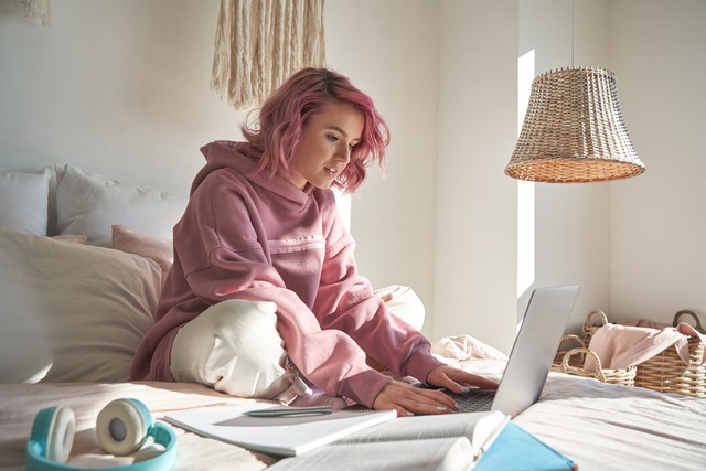 Young woman with pink hair sat on her bed reading from her laptop