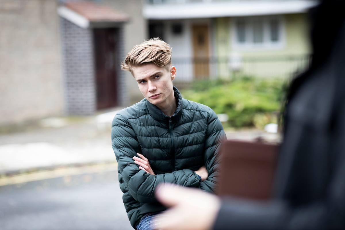 A young, blonde man is standing on the pavement with his arms crossed. He is thinking about his experience with friends and drugs. He is wearing a puffer jacket. This is a wide-angle image.