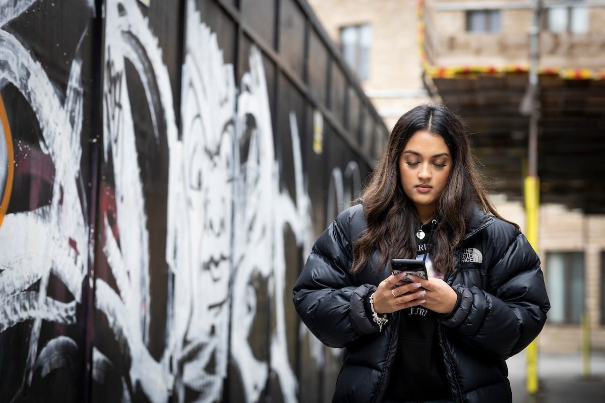 A young person wearing a black coat is standing on the street looking at their phone and googling: "why do people self harm?"