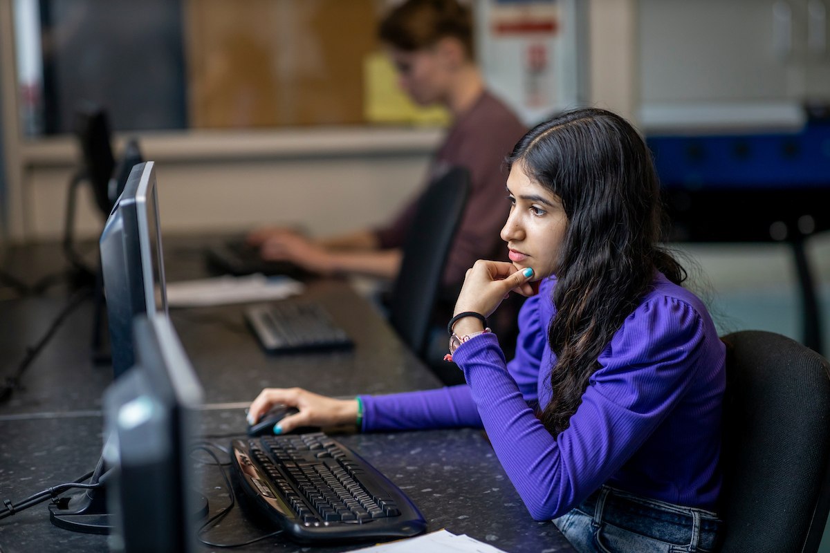 A young woman is sitting at a computer. She is researching online degrees. This is a wide-angle image.