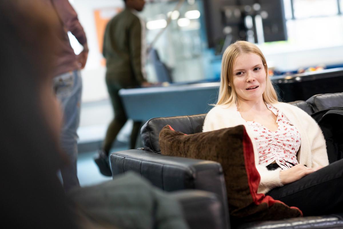 A young woman is sitting on a sofa. She is saying 'my girlfriend is depressed'. This is a wide-angle image.