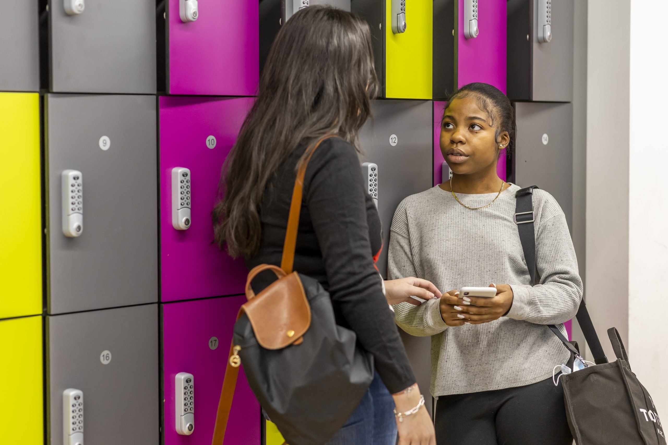 Two young people are standing in a school corridor, leaning against pink and grey lockers. One is wearing grey and the other is wearing black. They are talking about masturbation and wondering, what happens when you wank too much?