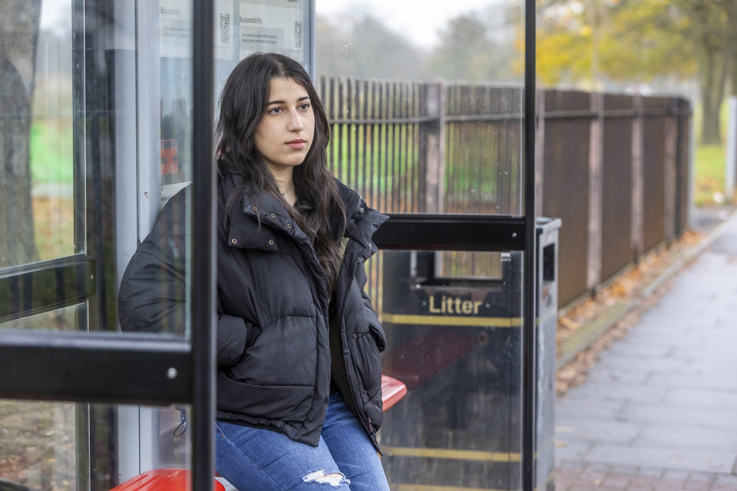 A young person is sitting at a bus stop wearing a black jacket, thinking about how to get support for sexual assault