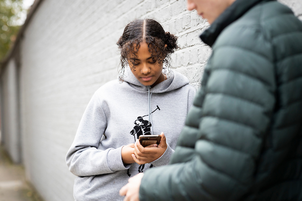 A young person is standing against a wall outside wearing a grey hoodie. They are looking down at their phone reading a guide to dealing with grief