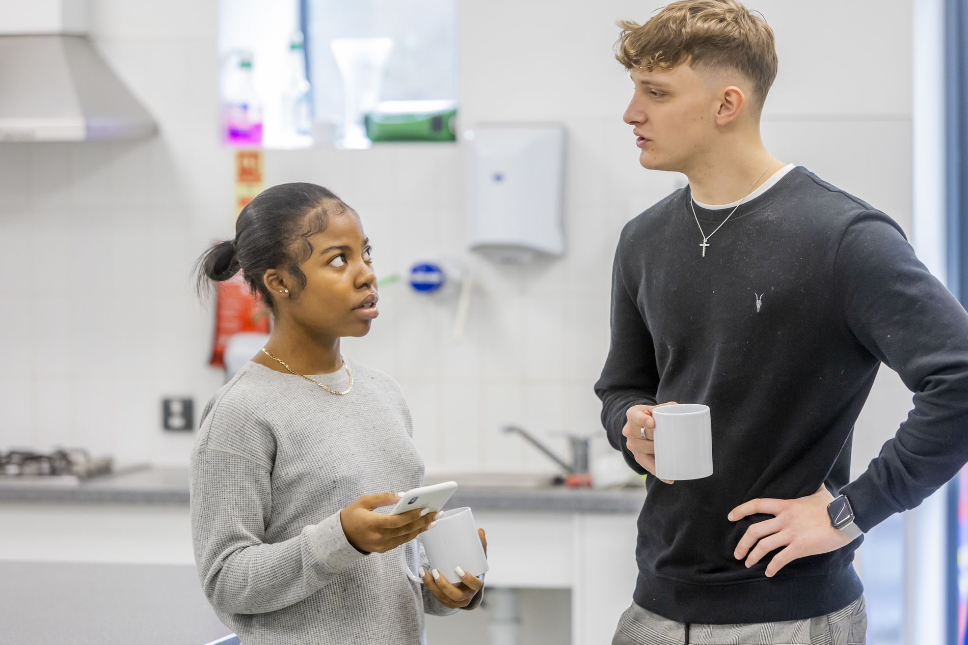 Two young people are talking about feeling lonely at Christmas. One is holding a mug and the other is holding their phone