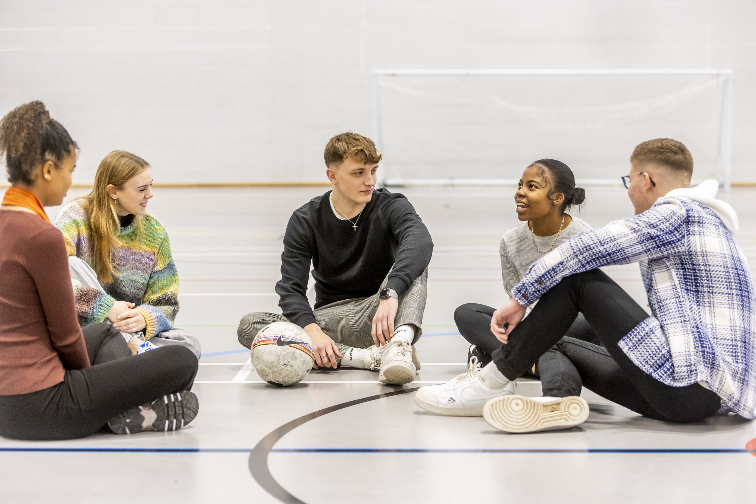 A group of young people are sitting on the floor of a school gym talking